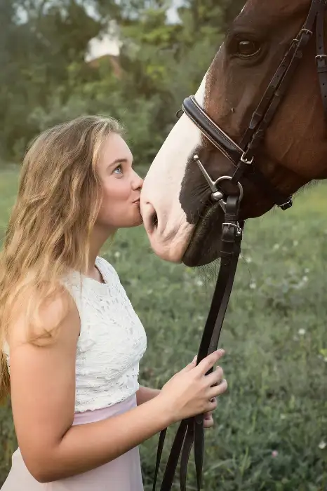 Junge Frau mit blondem Haar in weisser Bluse küsst ein braunes Pferd mit rosa Blässe auf die Nase